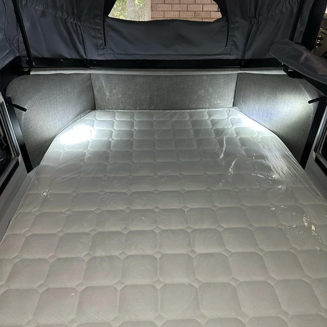Interior of a camper van bed area, with a white mattress on a flat surface, surrounded by gray fabric walls and a black framed opening.