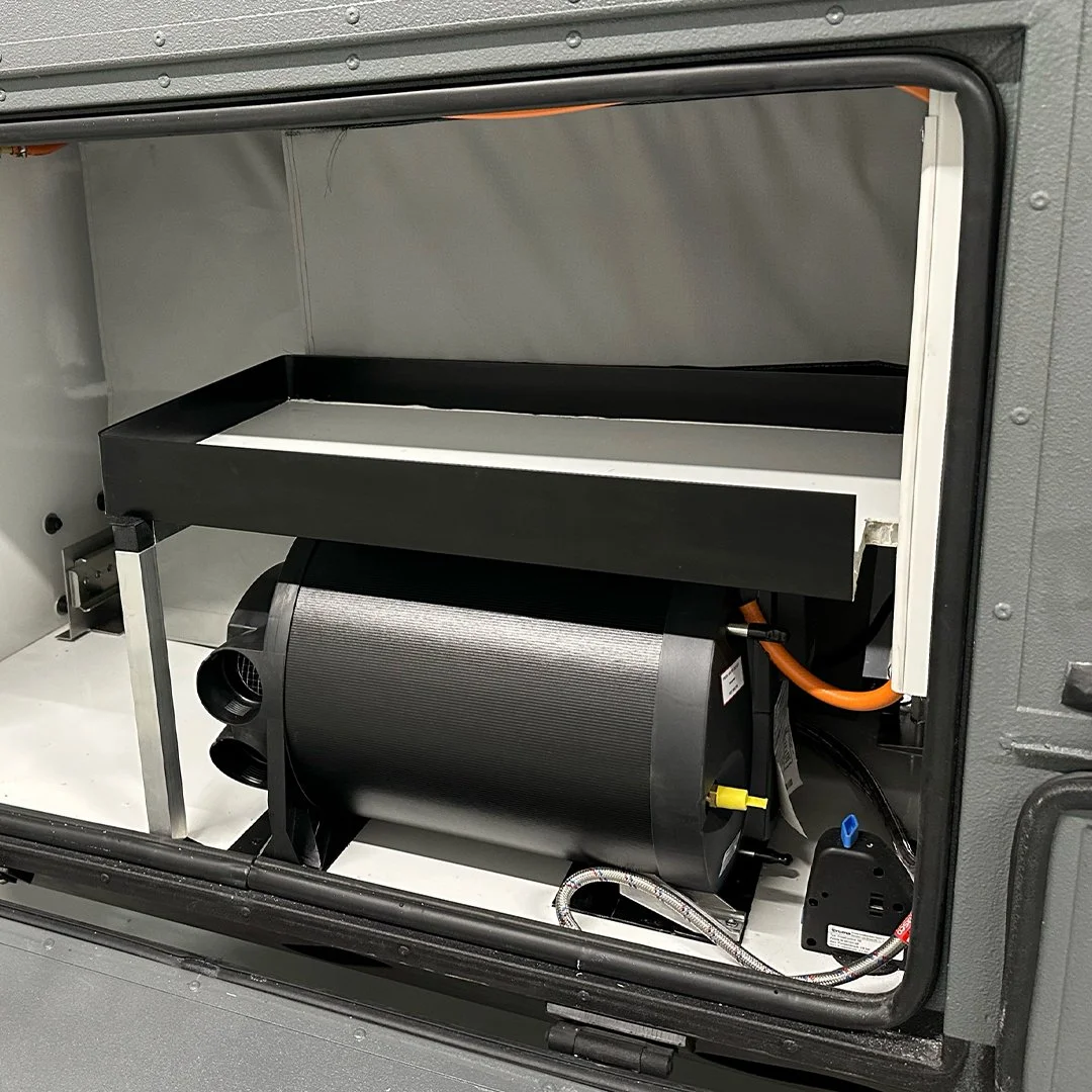 Interior view of a compartment containing a cylindrical black compressor with an orange power cord and a small black device, all inside a metallic enclosure.