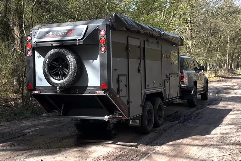 A gray BorderX off-road camper trailer attached to a pickup truck on a dirt trail surrounded by trees.