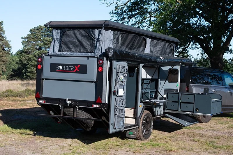A custom overland BorderX camper mounted on a truck with modular storage, an extending side deck, and a pop-up roof tent, parked on grassy terrain with trees in the background.