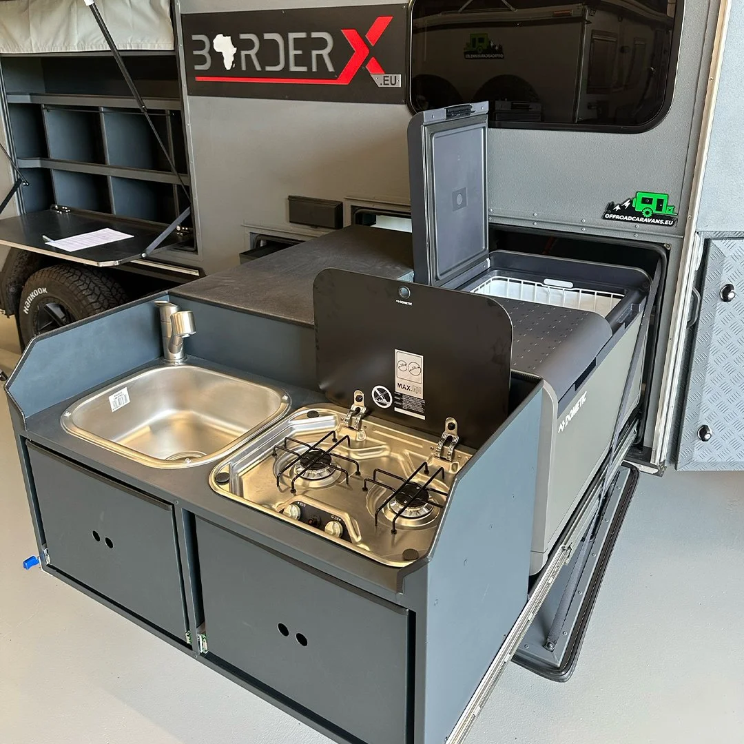 Camping trailer with compact outdoor kitchen featuring a stainless steel sink, two-burner gas stove, and refrigerator.