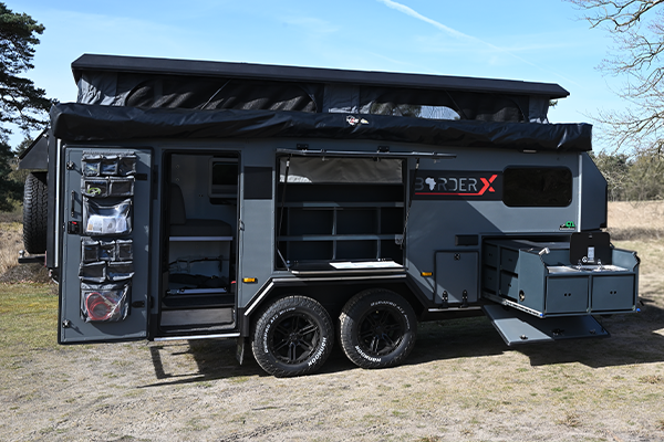 A black and gray BorderX off-road camper trailer with an extended slide-out kitchen and an awning. The trailer has rugged tires and various storage compartments.
