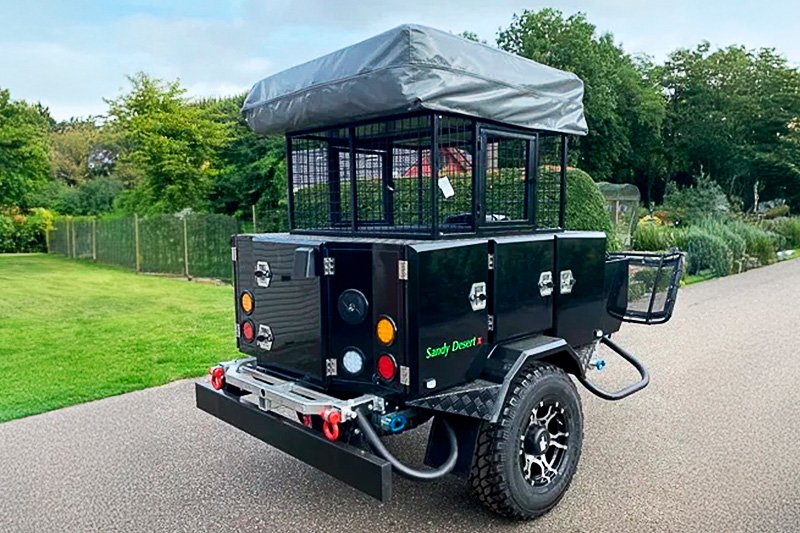 A compact off-road trailer with a rooftop tent and storage compartments, labeled 'Sandy Desert,' parked on a driveway with greenery in the background.