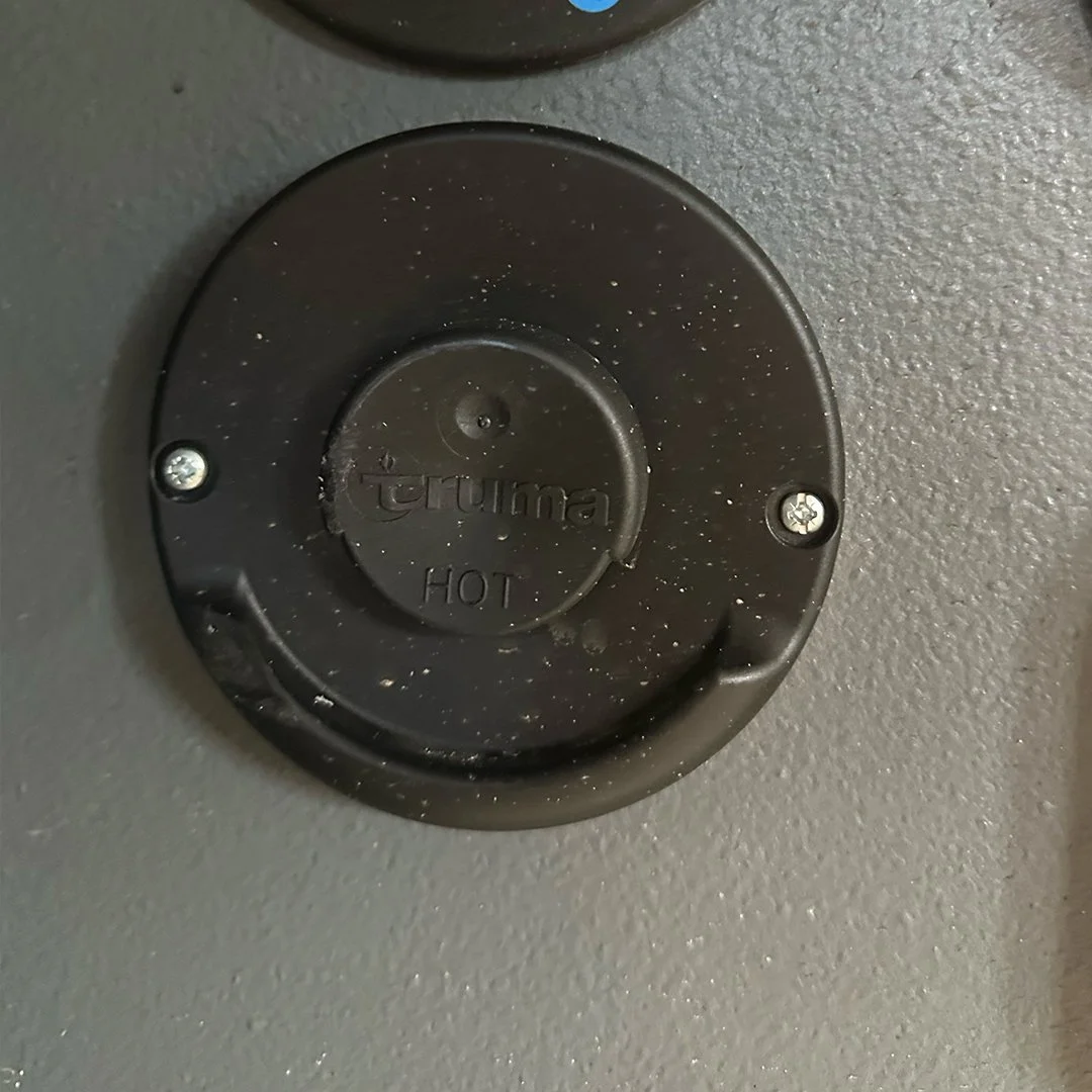 Close-up of a black circular cover labeled 'truma' and 'HOT' on a gray textured wall, secured with two screws.