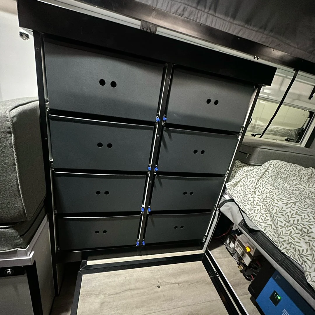 A black shelving unit with multiple compartments is placed inside a room, with a bed on the right side and a small section of a gray couch on the left.