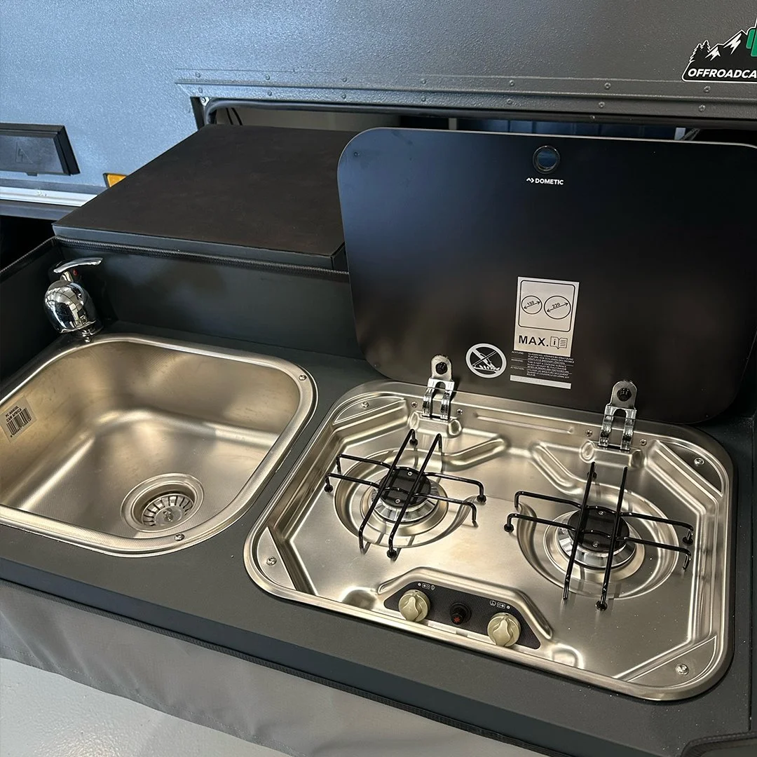 A stainless steel outdoor camping stove with two burners and a sink, built into a black countertop with a cover, on a gray surface.