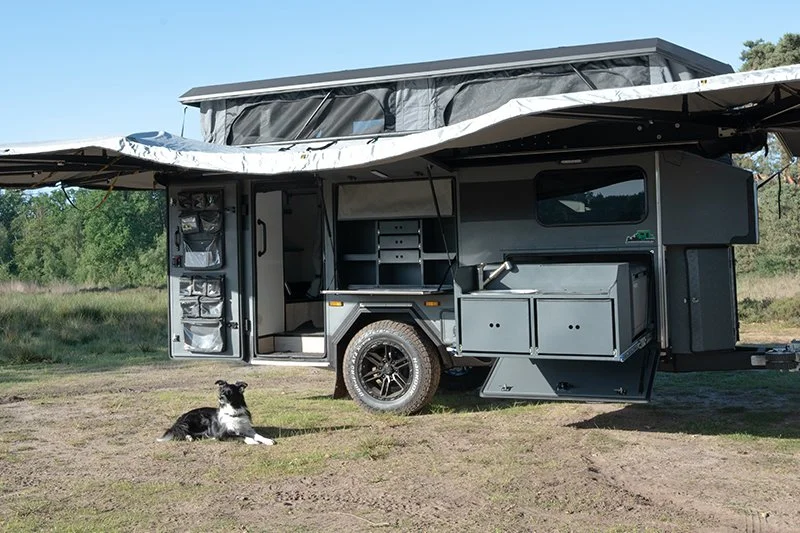 A black and white dog lying on the ground in front of a gray BorderX off-road camper trailer with a pop-up roof, in an outdoor grassy area.