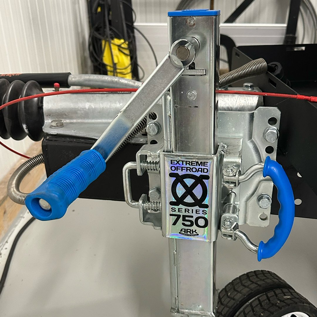 Close-up view of off-road vehicle steering components, including a metal framework labeled 'Extreme Offroad Series 750 Ard,' with blue grips on the steering wheel and other metal parts in a garage or workshop setting.