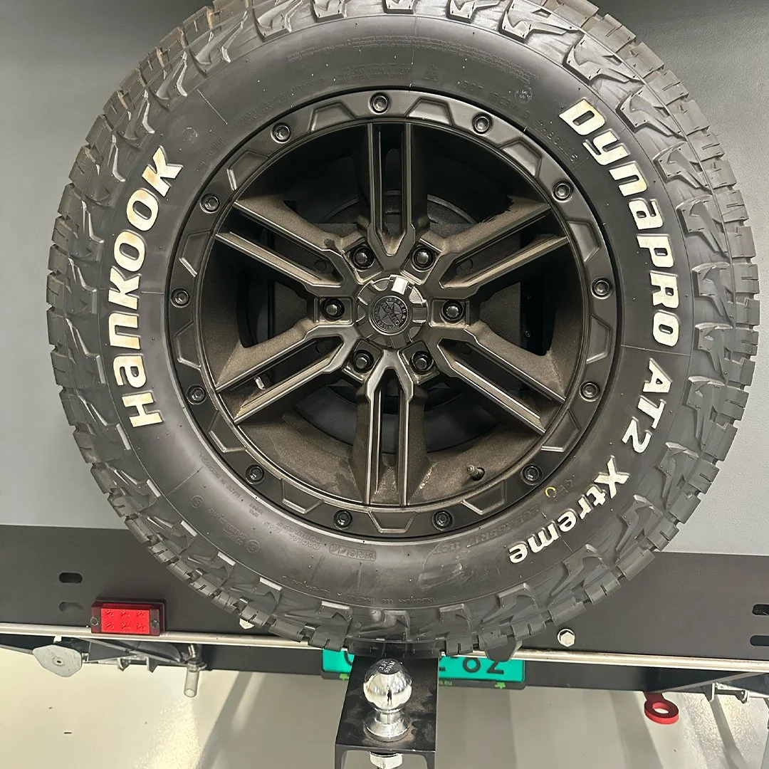 Close-up of a vehicle wheel featuring a Hankook Dynapro AT2 tire and a black, multi-spoke rim mounted on a trailer or vehicle. The tire has rugged tread pattern and white lettering.