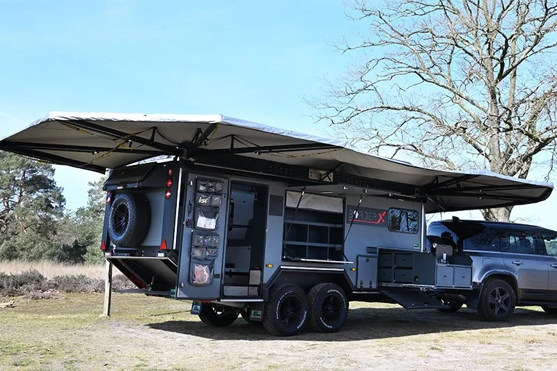 BorderX Off-road camper trailer attached to a vehicle with extended awnings, set up outdoors in a grassy area with trees and clear blue sky.