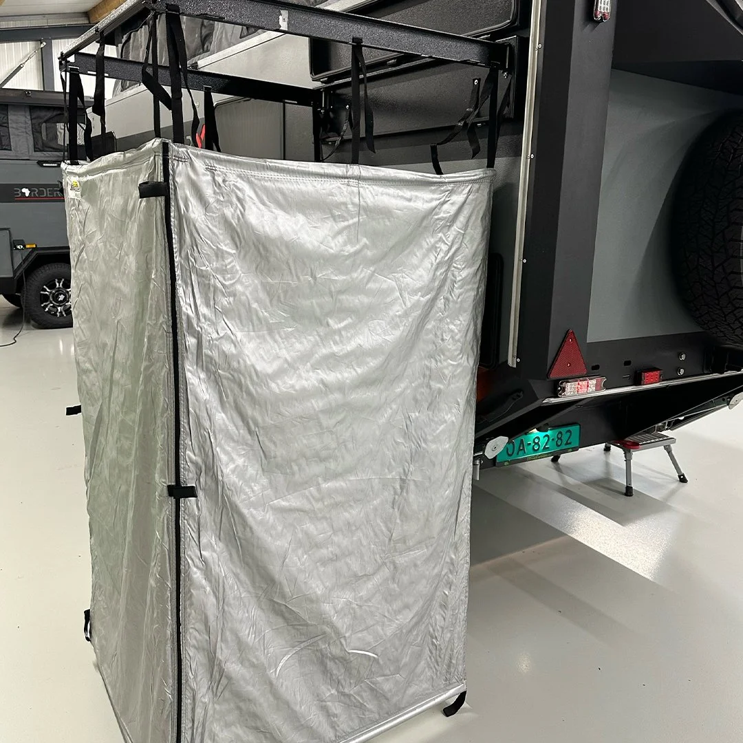A portable folding tent or shelter with a silver reflective surface, mounted on black straps attached to a vehicle or trailer in an indoor setting.