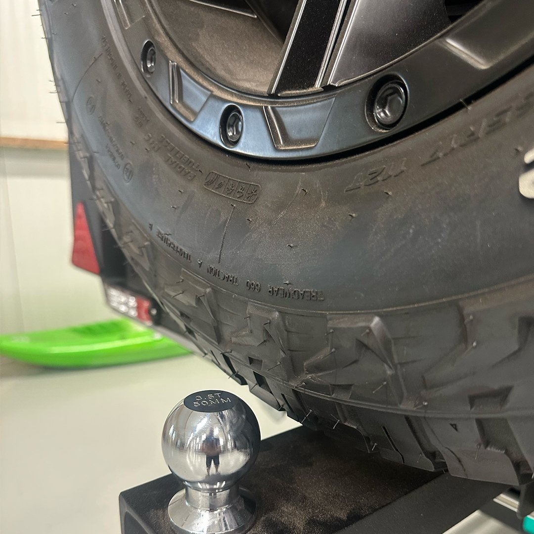 A close-up of a vehicle tire mounted on a wheel, with a shiny metallic weight labeled '3.5 FT 50MM' positioned near the tire for balancing, all on a black surface.