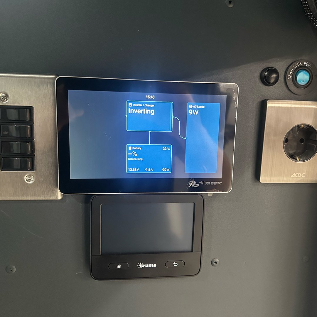 A digital monitor displaying an energy management interface, showing inverter status, battery temperature, and power details, mounted on a gray panel with a blue power socket and black switches.