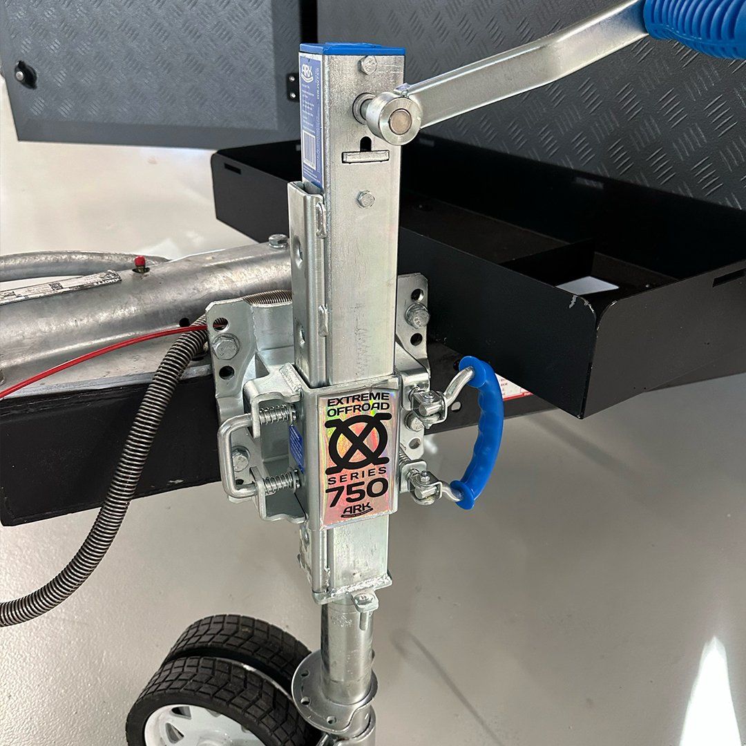 Close-up of a trailer coupling device with a safety latch and label, attached to a black trailer frame, with a wheel at the bottom.