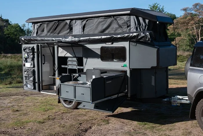 A converted camper trailer with outdoor kitchen setup, fold-out counter, and storage compartments, parked on grass with trees in the background.