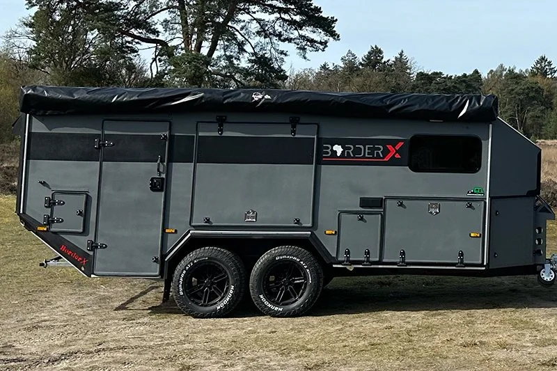 A gray BorderX off-road camper trailer with multiple storage compartments, black wheels, and a black cover on top, parked on grass with trees in the background.