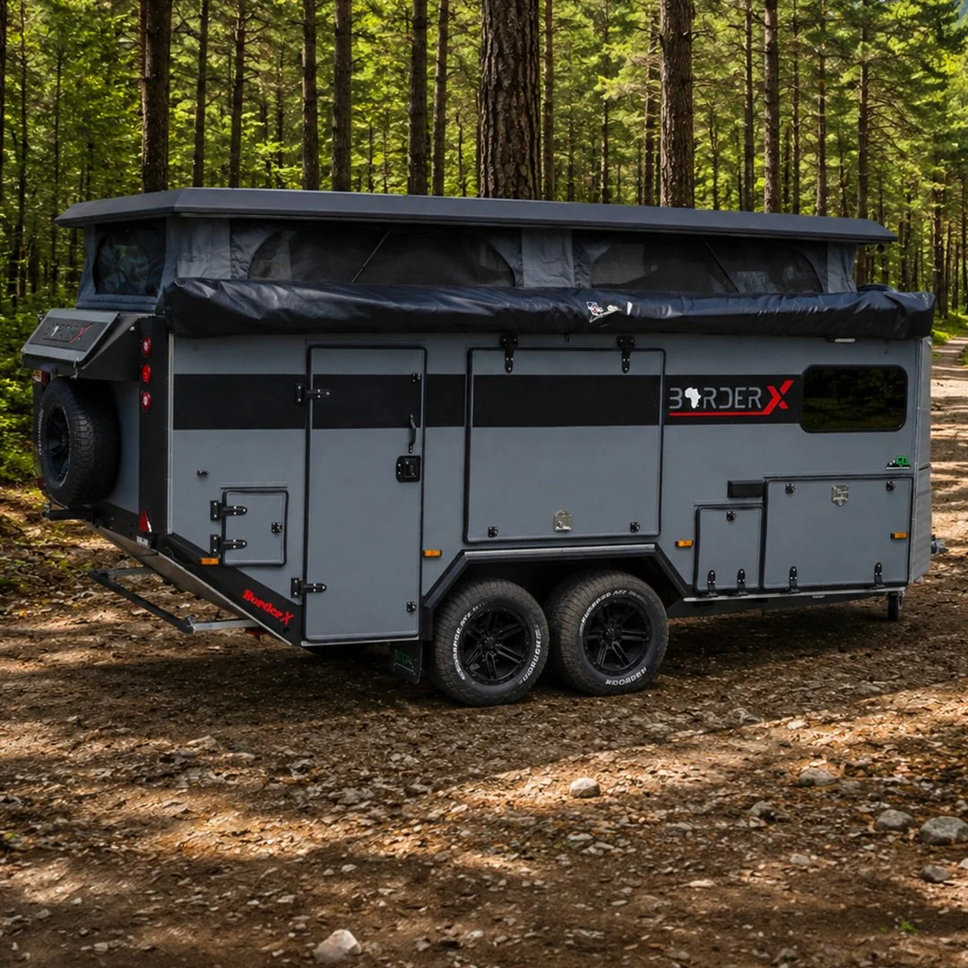 A compact, gray off-road BorderX camper trailer with black and red accents, parked in a forested area with dirt ground, surrounded by tall green trees.