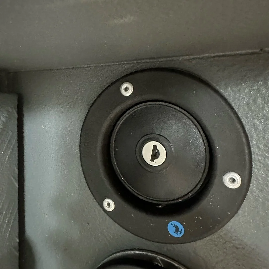 Close-up of a black speaker or camera lens with a central keyhole-like slot surrounded by a black circular mount, affixed to a textured black surface with four screws.
