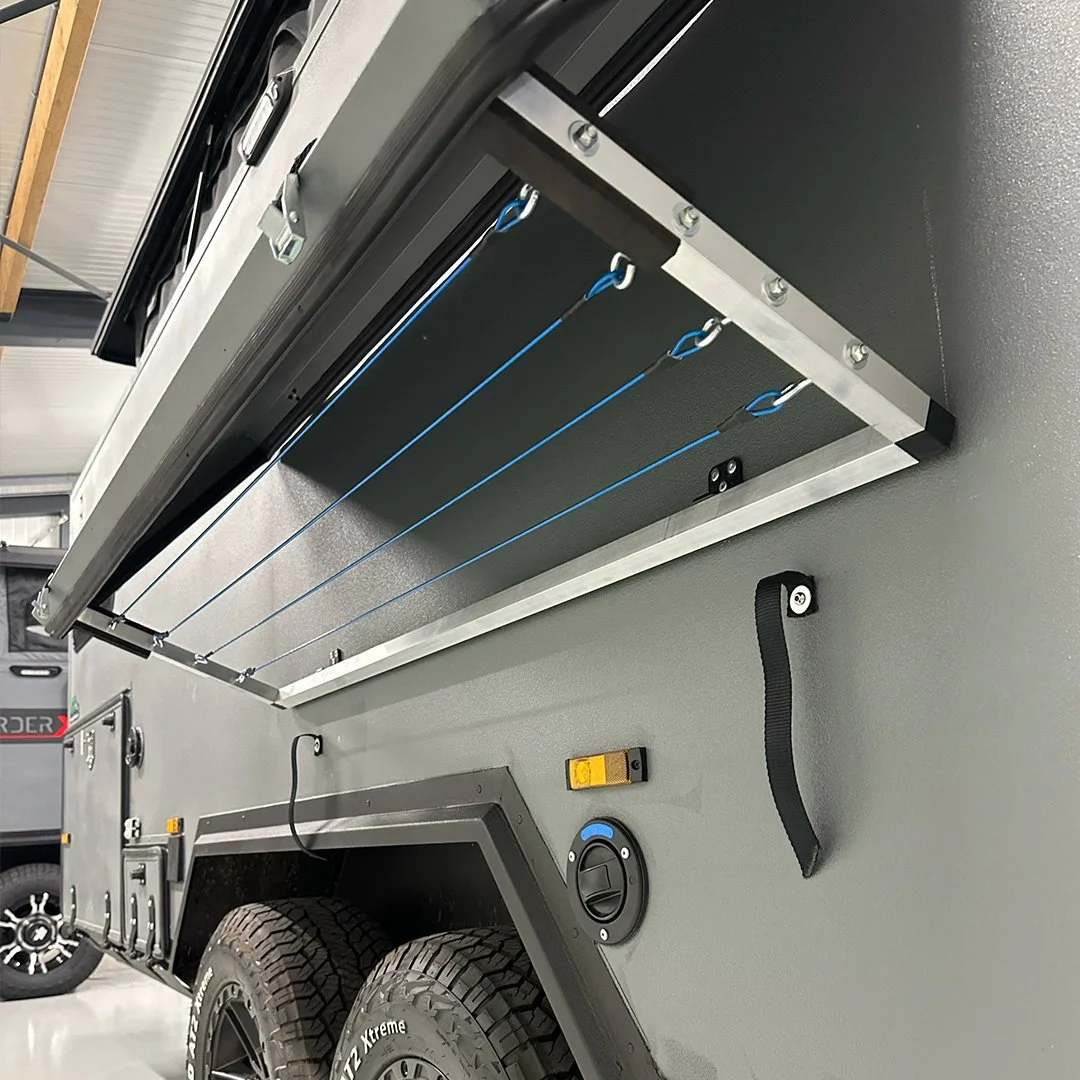 Close-up of a rugged off-road trailer with four blue high-tensile cords attached to hooks, mounted on the side of the trailer. The trailer has large off-road tires and a panel with a dial, a yellow switch, and a black strap.