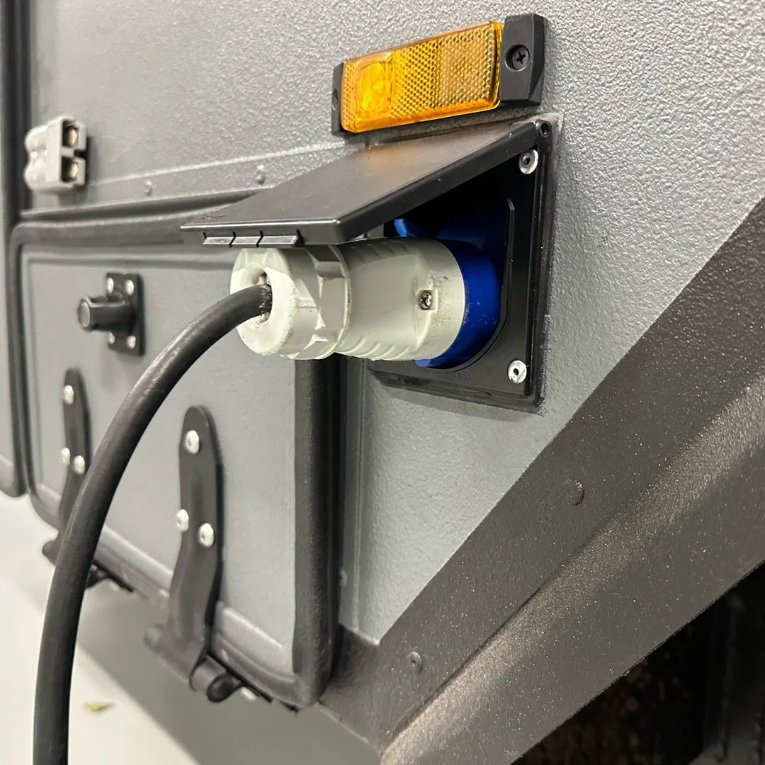 Close-up view of an electrical outlet with a plugged-in black cable mounted on a gray industrial or commercial equipment surface, with a small orange warning light on top.