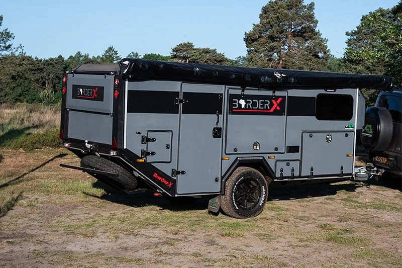 Gray off-road BorderX amper trailer with black accents parked on grass, attached to a black vehicle, with trees in the background.