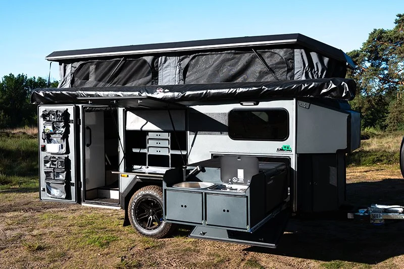 A modern, gray and black BorderX camping trailer with an outdoor kitchen and storage compartments, parked outdoors on a grassy area with trees in the background.