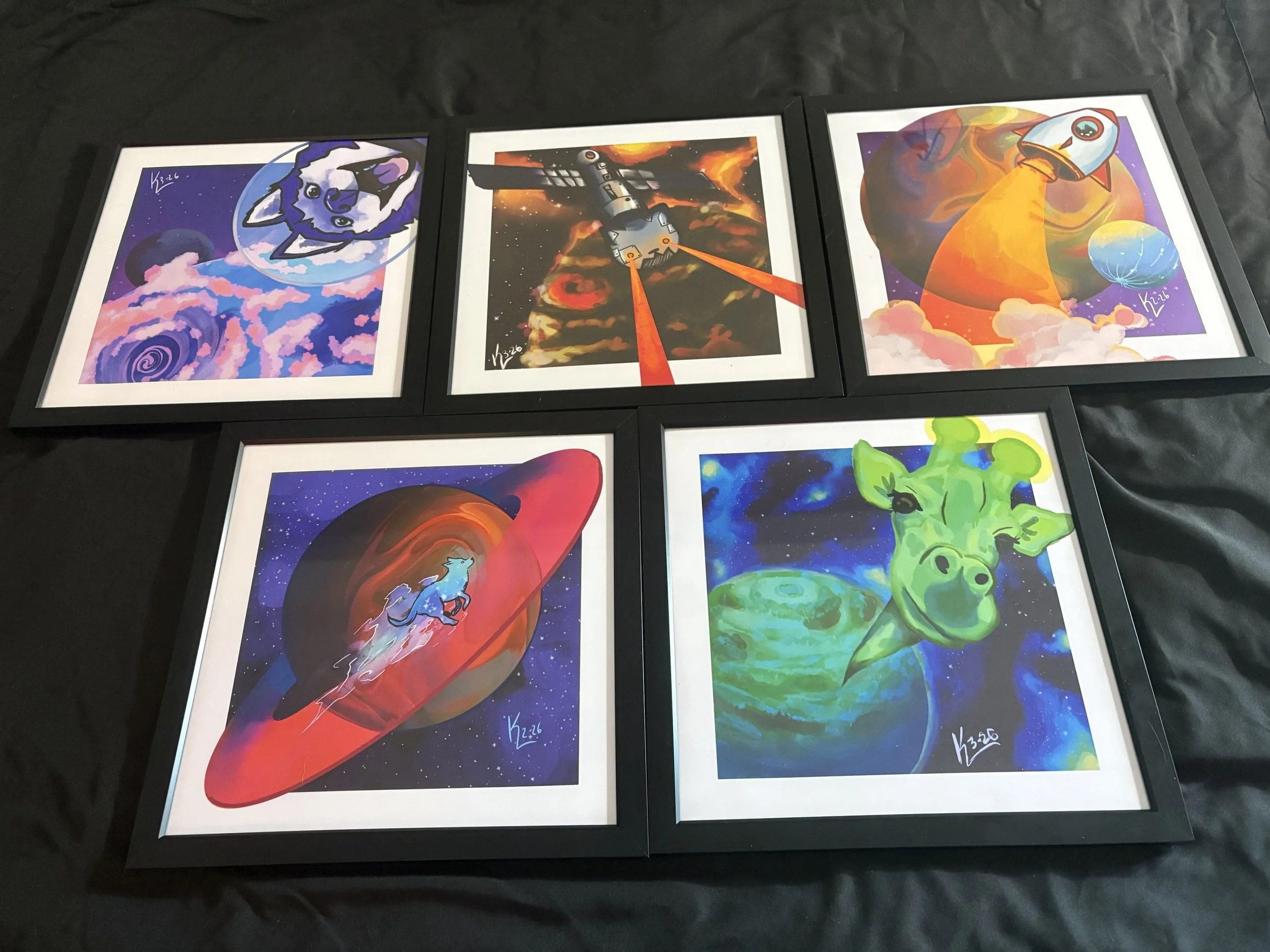 Five colorful space-themed digital art prints in black frames displayed on a black surface. The artworks include a husky dog in space, a spaceship firing lasers at planets, a rocket launching from a planet, a fox running through space around a planet, and a smiling cow floating in space near a planet.