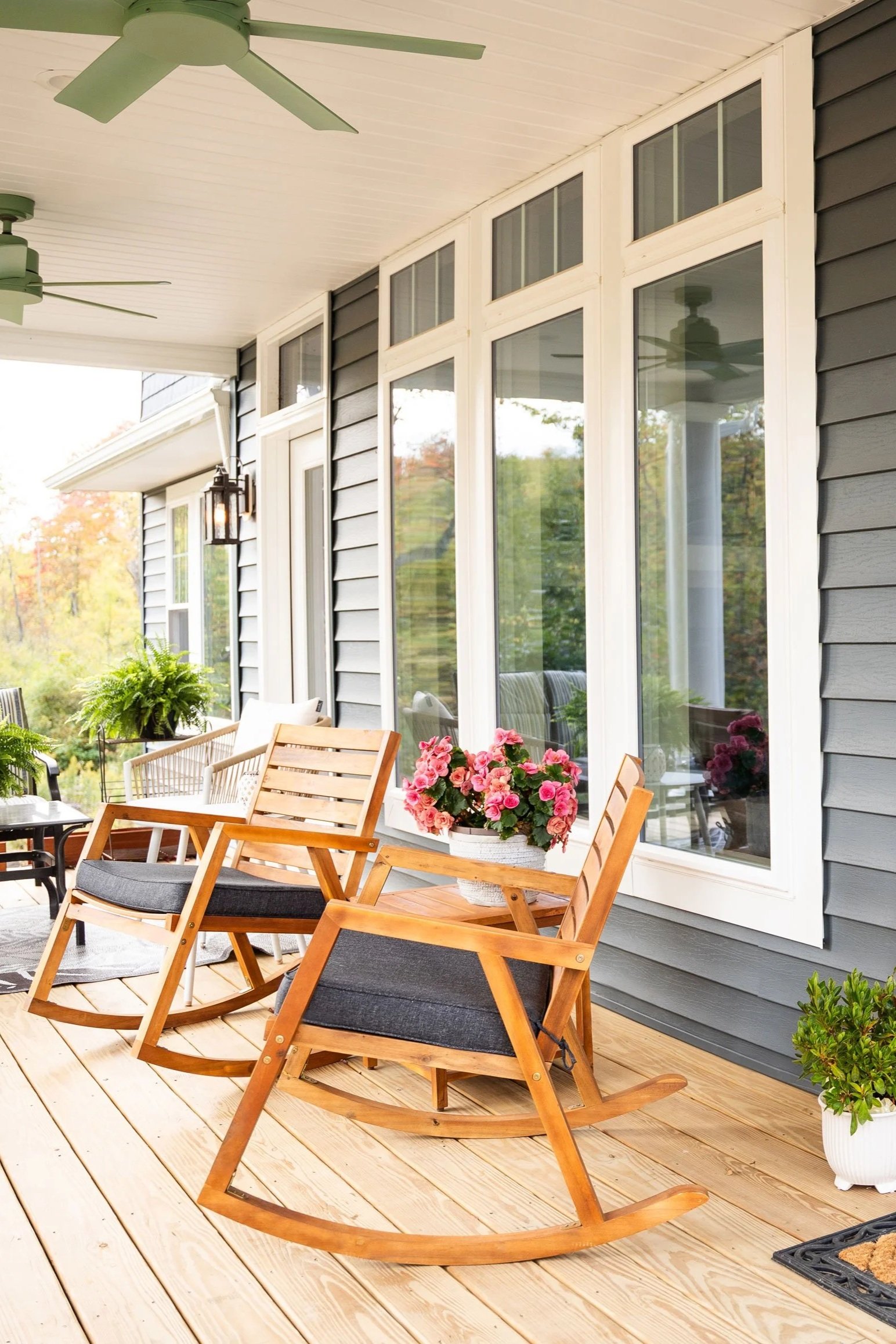 A porch with wooden flooring, gray siding, large windows, a ceiling fan, a hanging lantern, and outdoor furniture including a rocking chair, chairs with gray cushions, and potted pink flowers.