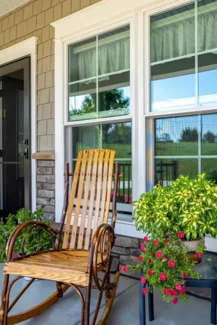 A wooden rocking chair on a porch next to a large window with white framing, surrounded by green plants and pink flowers in a pot.