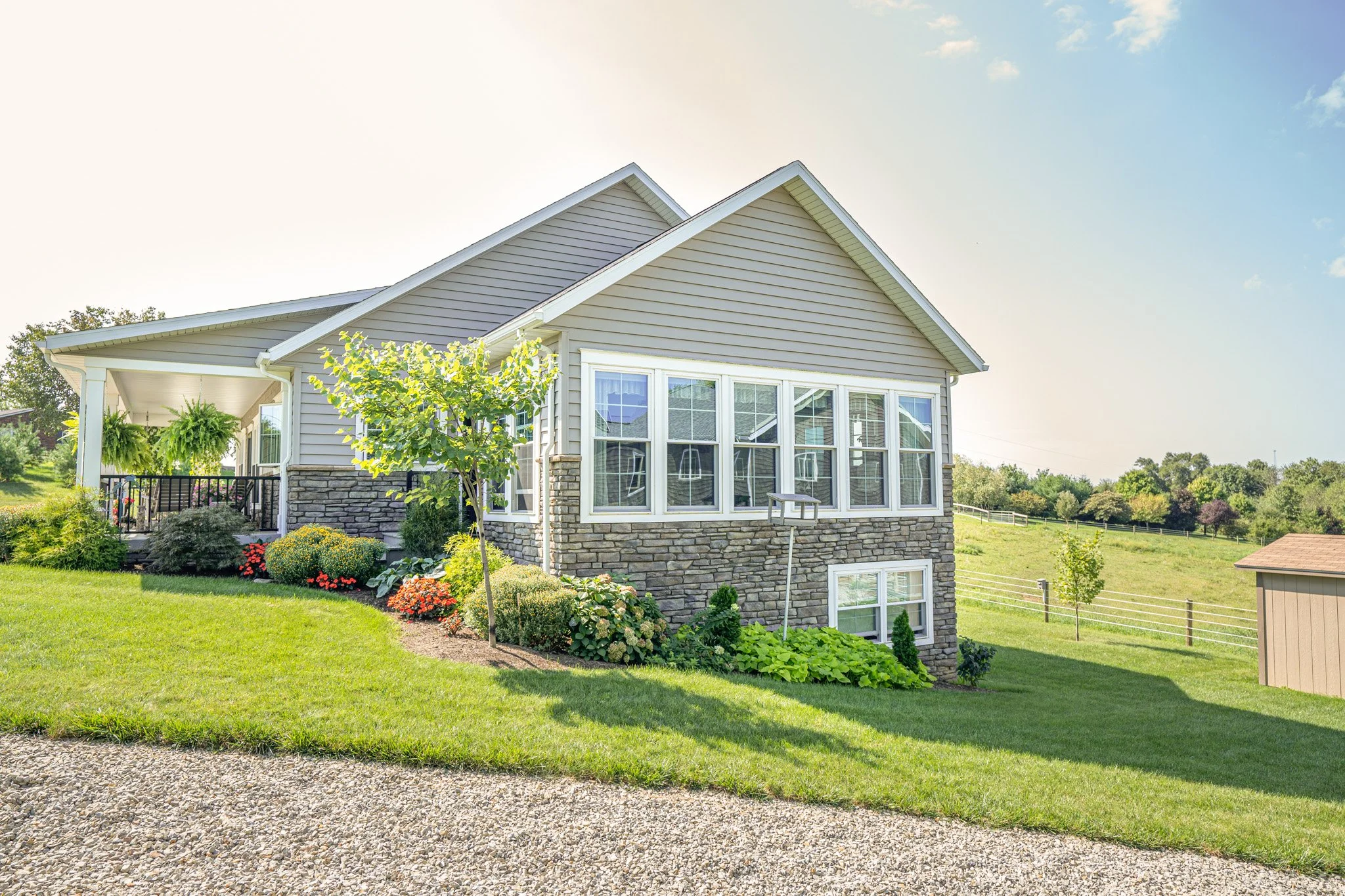 Front view of a house with beige siding, large windows, a stone foundation, and a porch with hanging plants, surrounded by a well-maintained lawn and garden, under a partly cloudy sky.