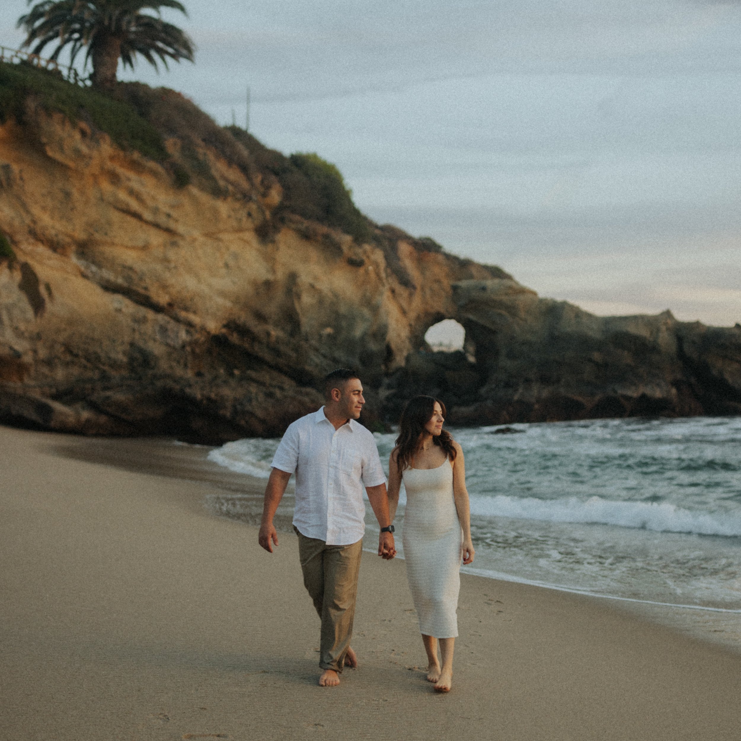 A couple holding hands while walking barefoot on a sandy beach, with rocky cliffs and ocean waves in the background. The scene is set during a calm, overcast day.