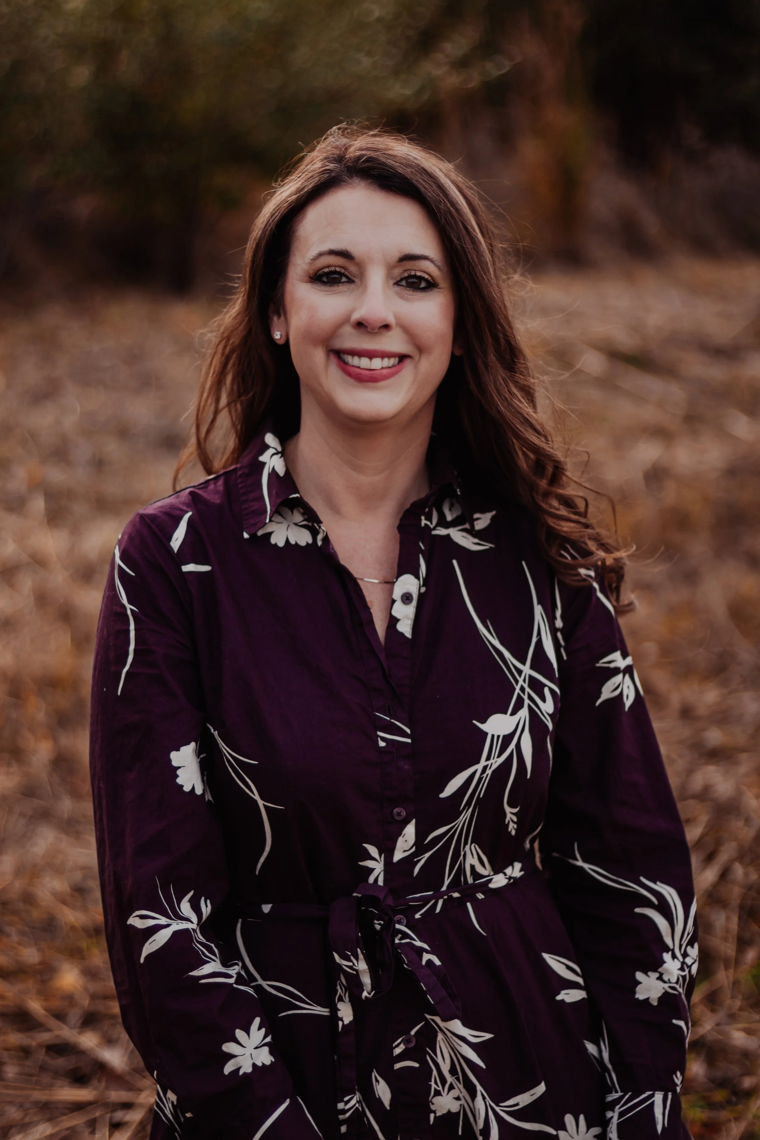 A woman with long, wavy brown hair, smiling, standing outdoors in front of a blurred autumn background. She is wearing a dark purple dress with a white floral pattern and a tied waist.