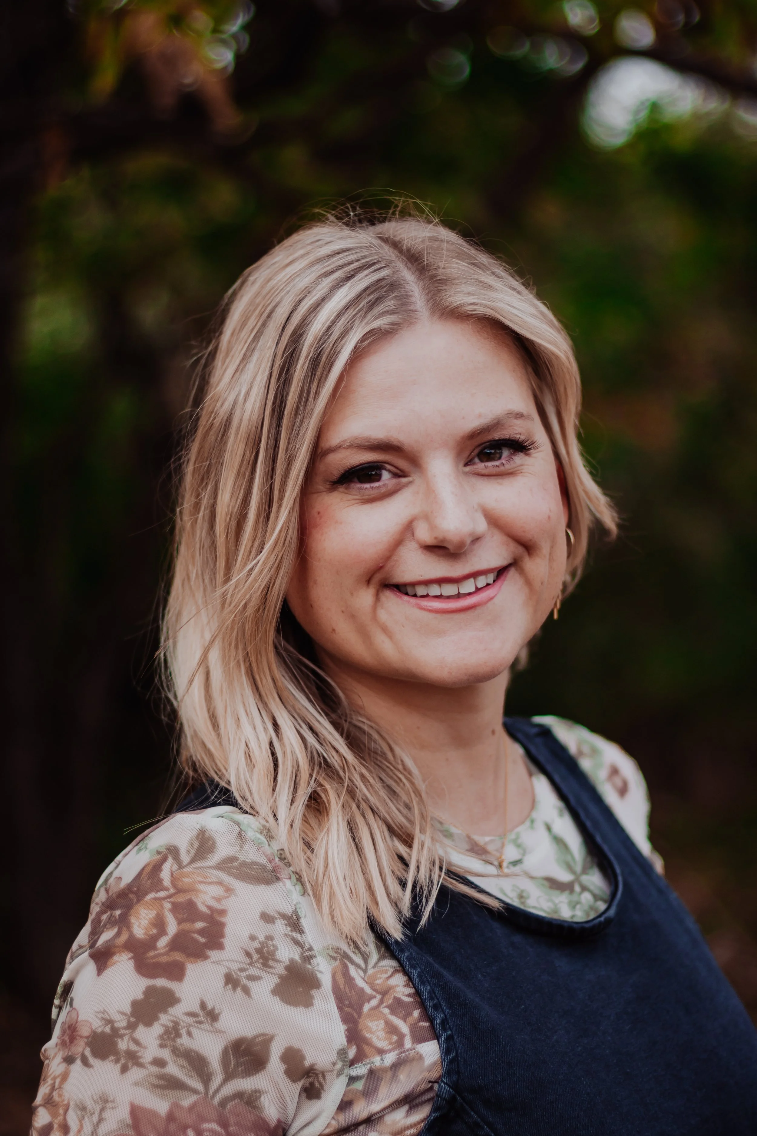 A smiling young woman with blonde hair wearing a floral-patterned top and a dark sleeveless overlay, standing outdoors with green foliage in the background.