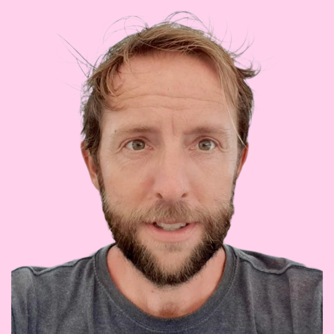 A man with light skin, brown hair, and a beard takes a selfie against a pink background.