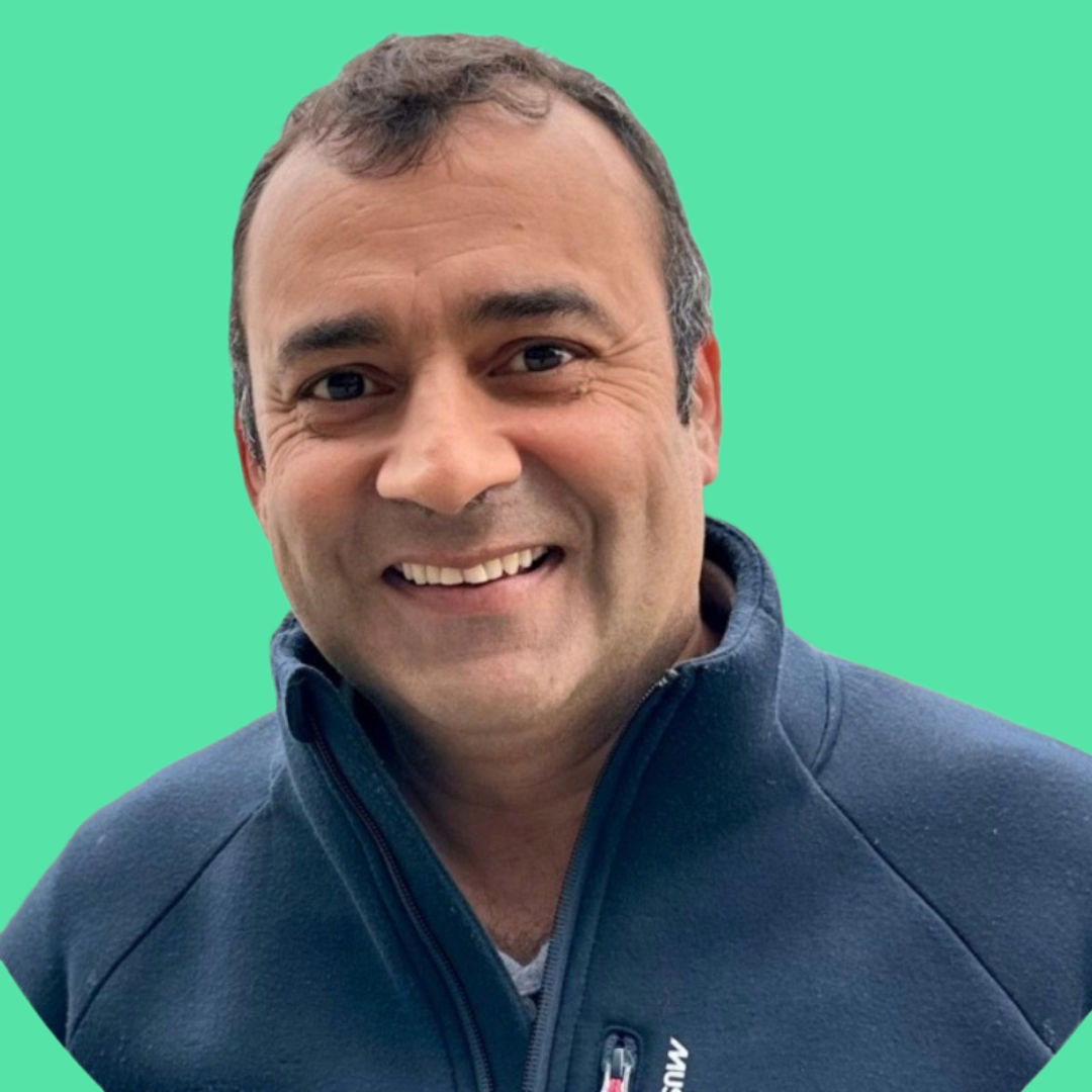 Close-up of a smiling man with short brown hair, wearing a navy blue jacket, with a solid light turquoise background.