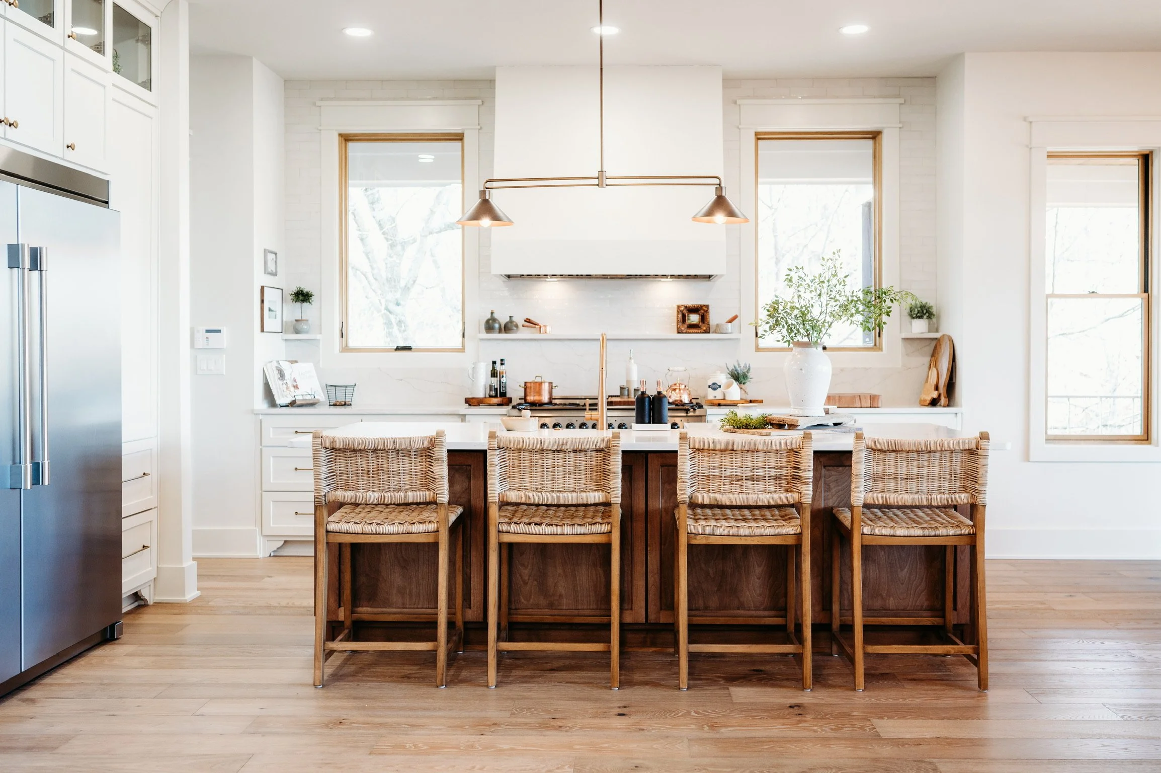 White modern kitchen with wooden island, four woven chairs, large windows, plants, and minimal decor.