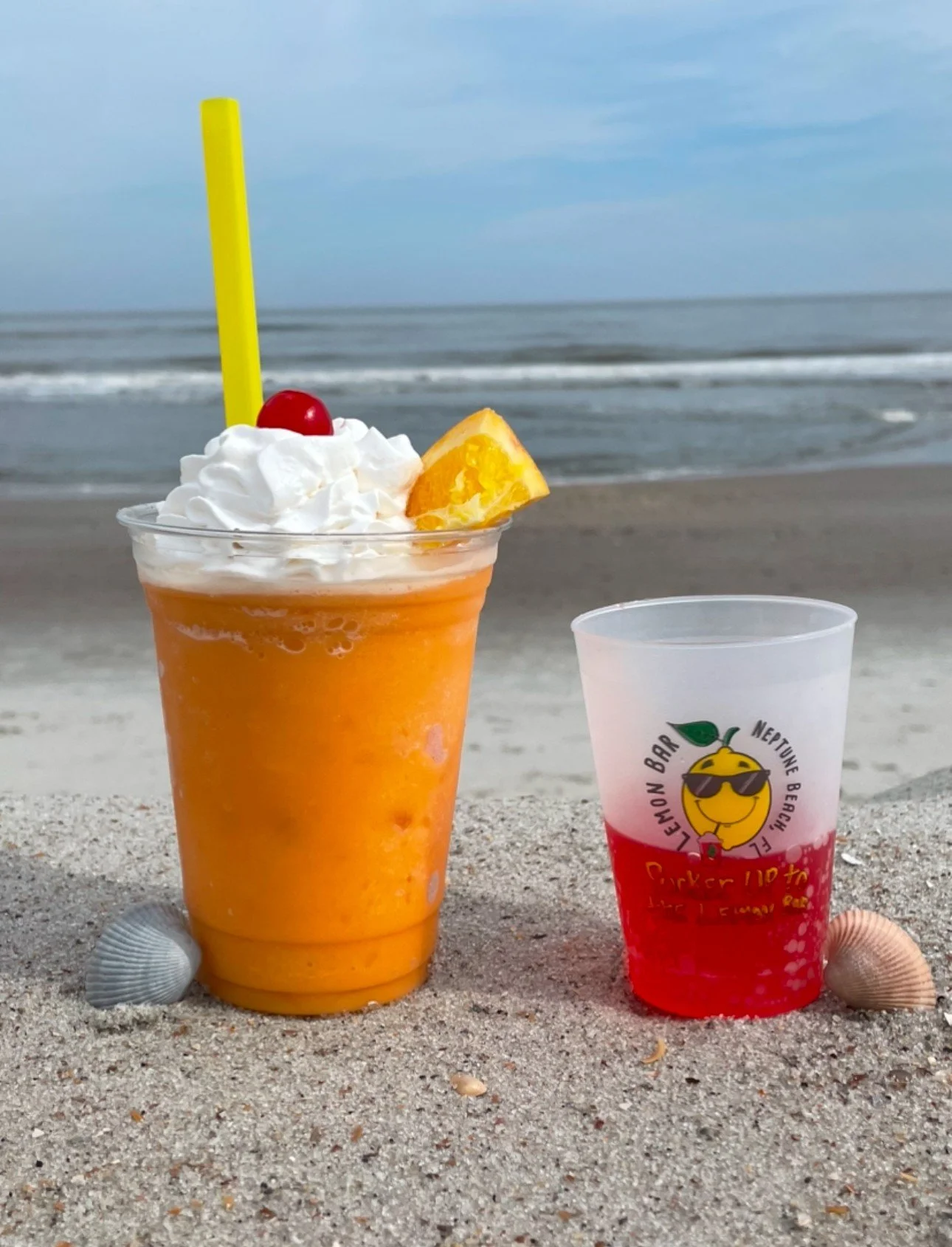 A tropical orange drink topped with whipped cream, a cherry, and a lemon wedge, on a sandy beach with seashells, with a second red and white beverage container nearby, and the ocean in the background.