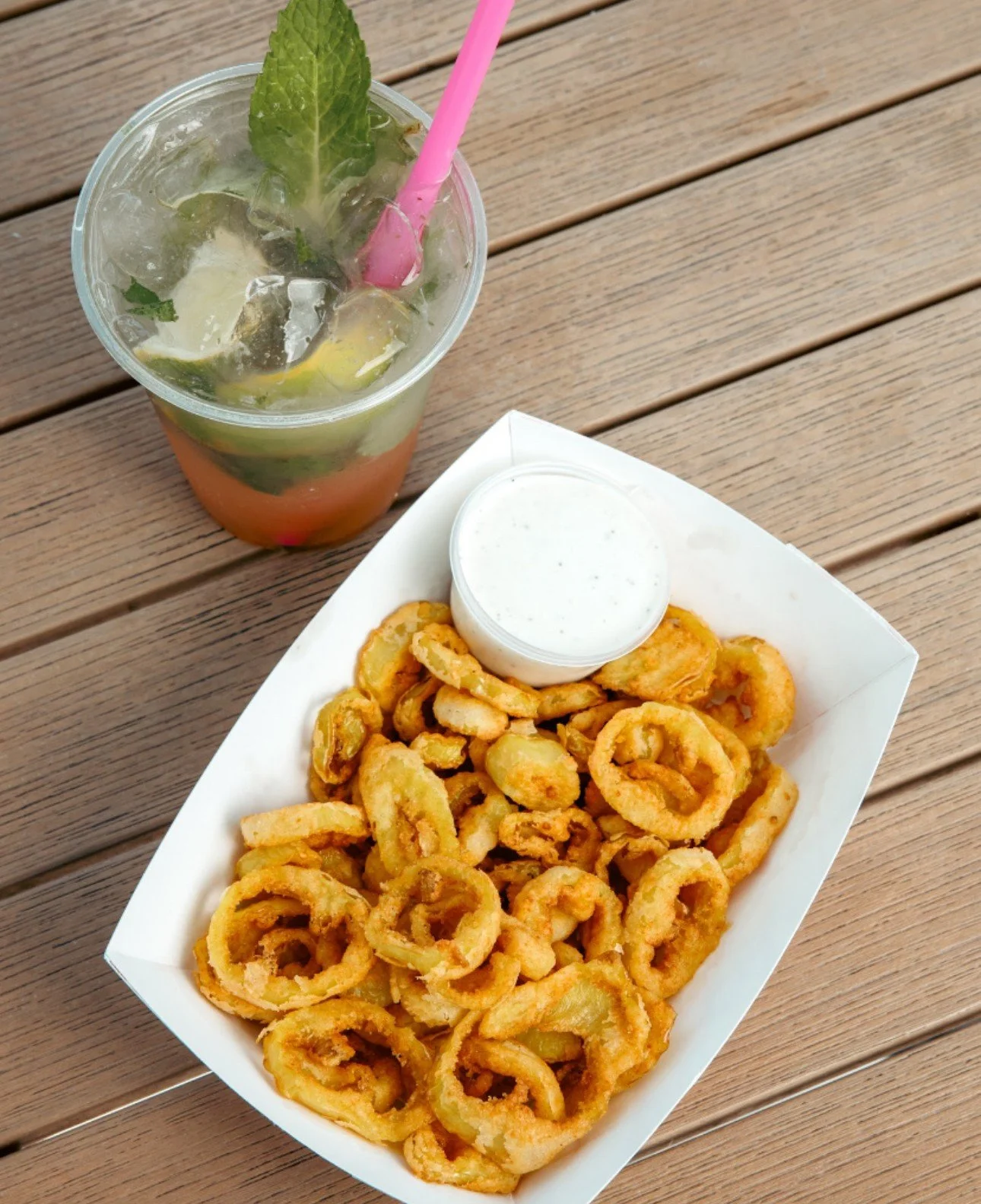 A tray of fried calamari with a small cup of white dipping sauce accompanied by a glass of iced lemon mint drink with ice, lemon wedges, mint leaves, and a pink straw.