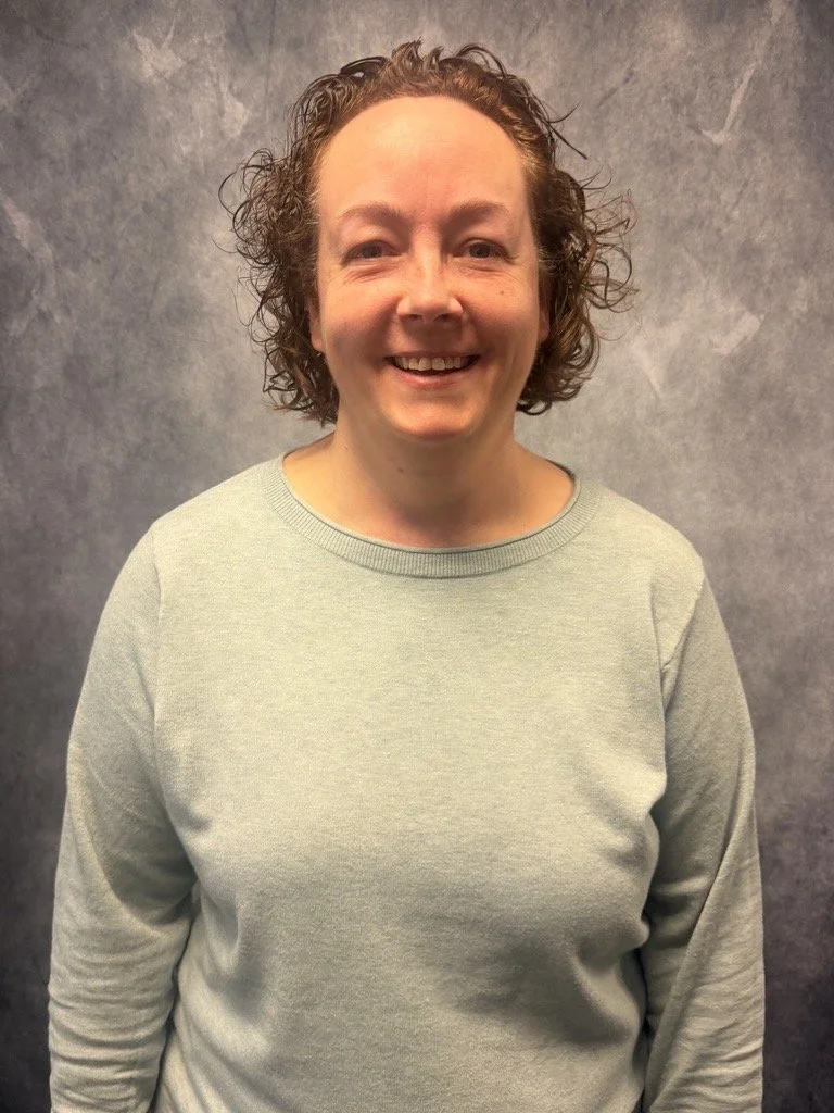 The image contains a white woman with curly brown hair in front of a gray background.