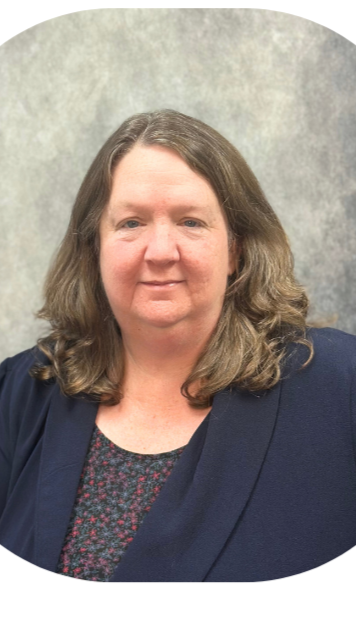 A woman with shoulder-length wavy brown hair and light skin, wearing a dark blazer and a patterned top, standing against a neutral background.