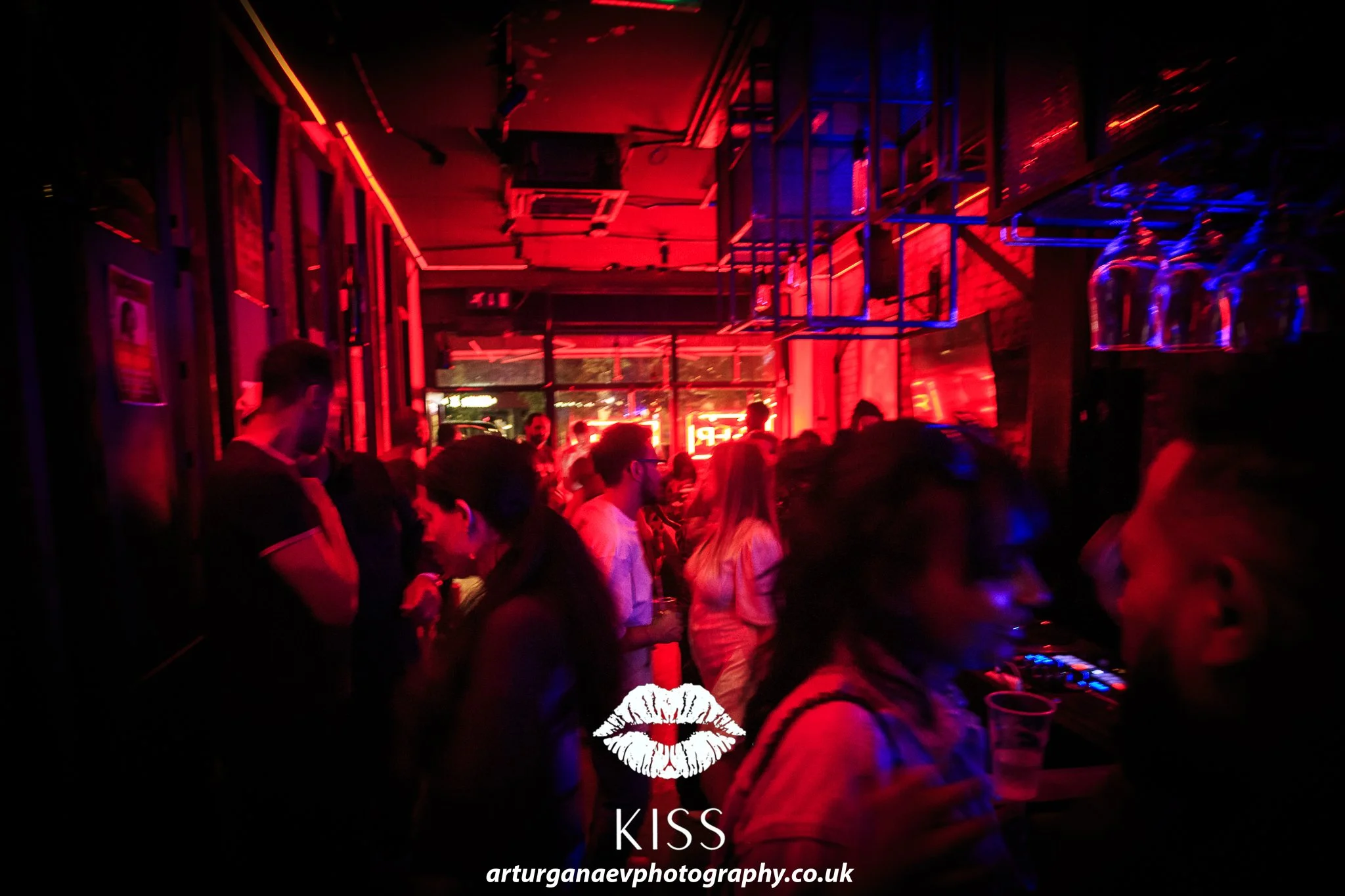 People socializing inside a bar with red and blue neon lighting. Some individuals are holding drinks, and glasses are hanging from a rack overhead.