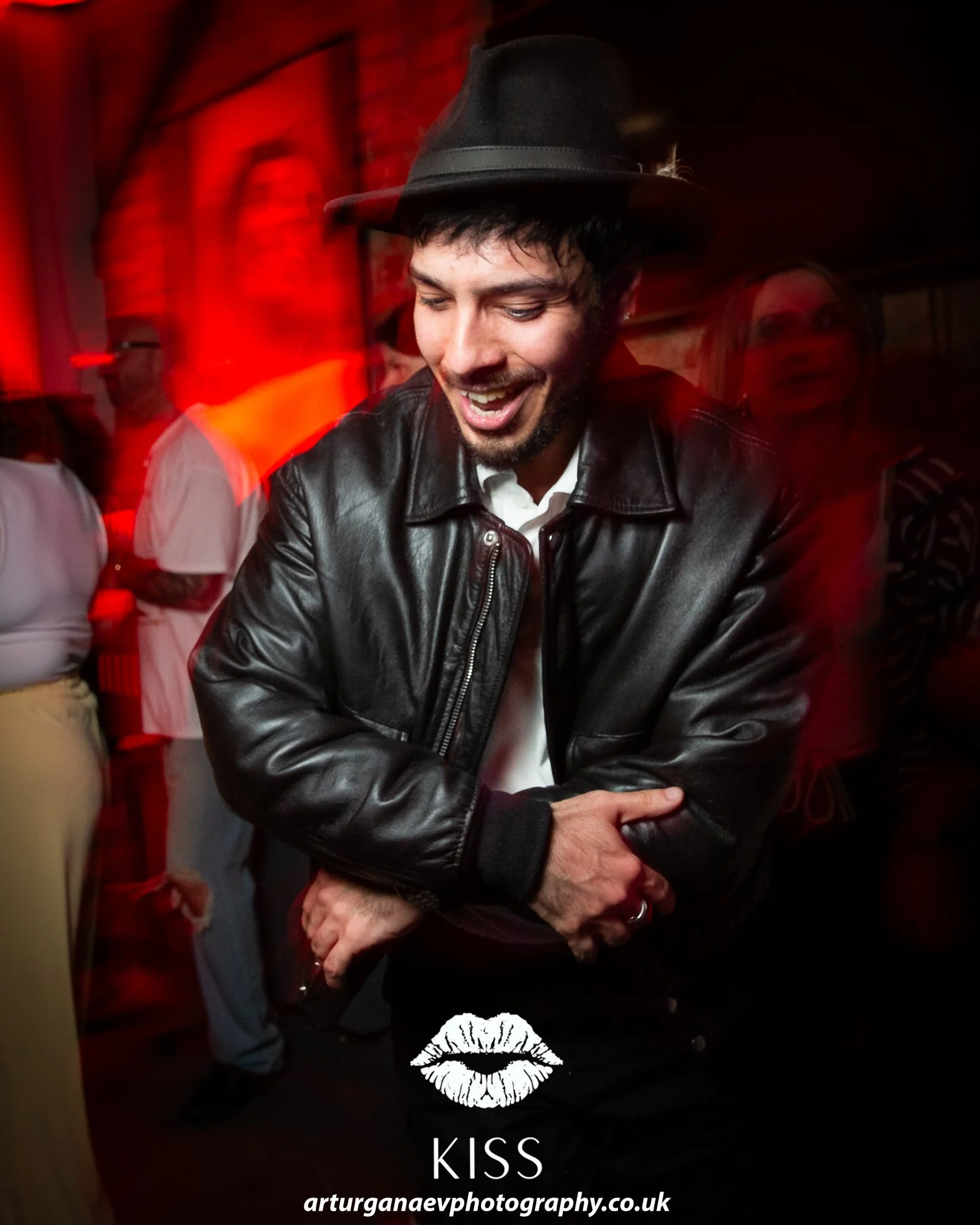 A man wearing a black leather jacket and black hat is smiling in a dimly lit bar or club with red lighting. Behind him, people are socializing. At the bottom of the image are a graphic of lips and the word 'KISS', along with a URL for a photography w