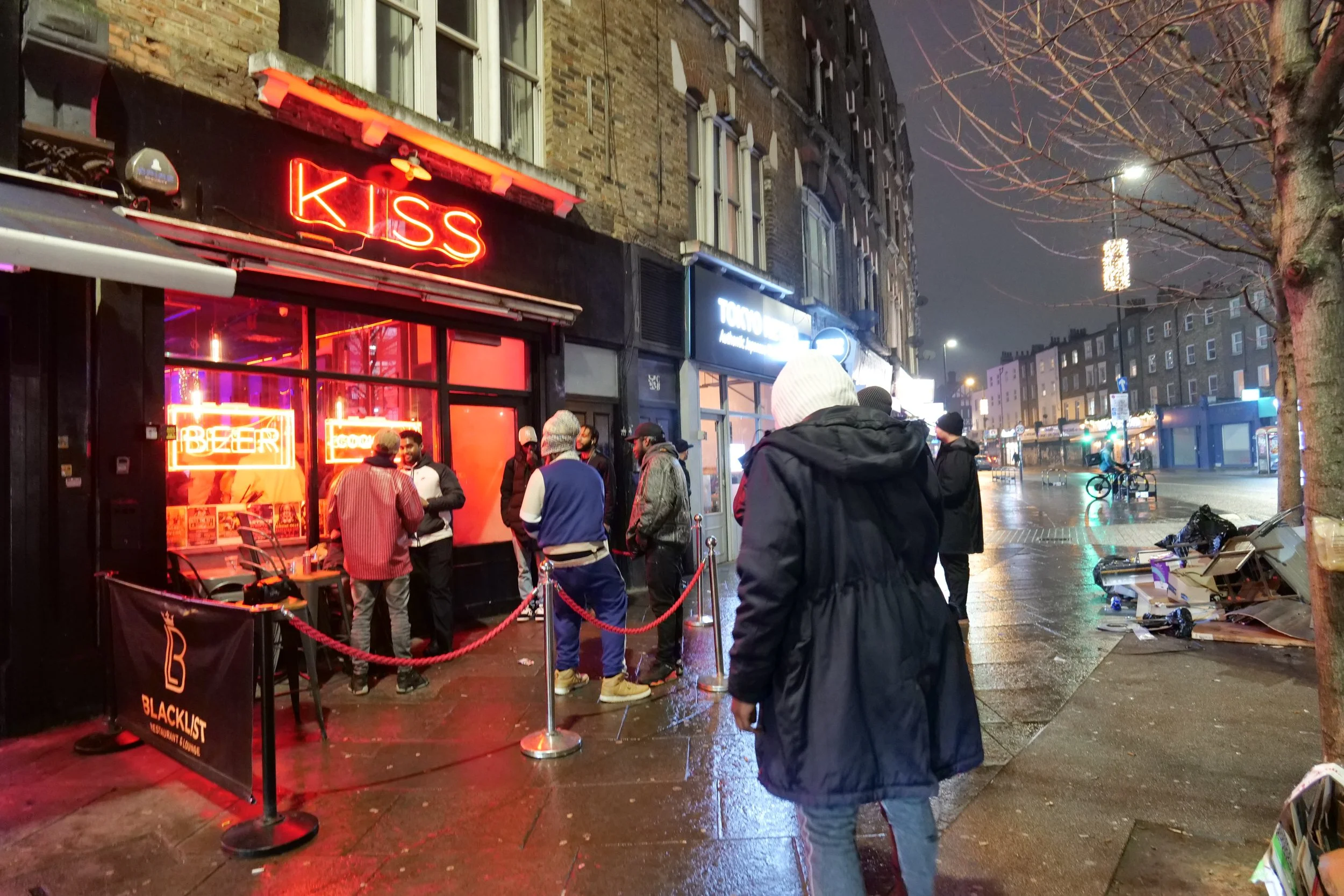 People standing outside a bar on a wet city street at night with neon signs, including a red "KISS" sign and a "BEER" sign inside the window, some people wearing jackets and hats, and trees with bare branches.