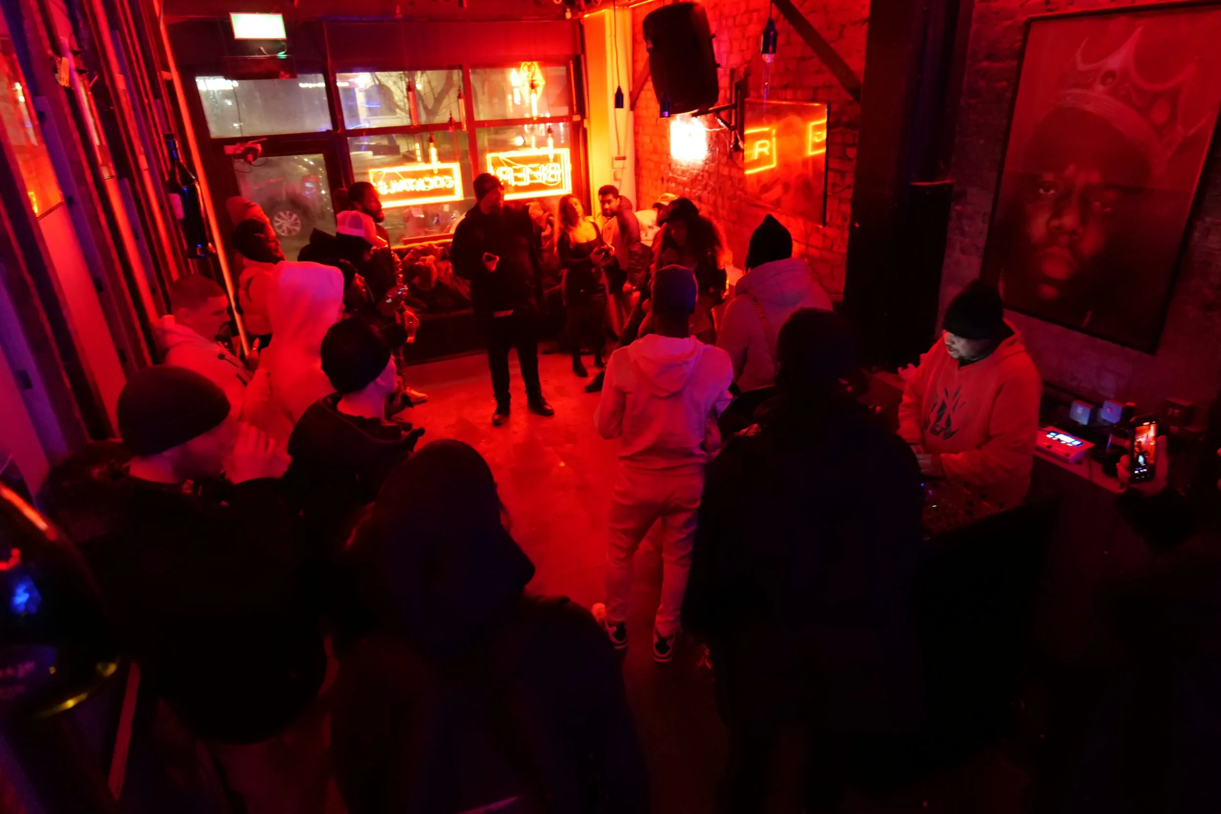 A crowded indoor space with people socializing and standing in front of a brick wall. The room has a red neon sign that says 'BEER' and 'WINES', and there are fluorescent lights and artwork on the walls. Some people are sitting, some are standing, an