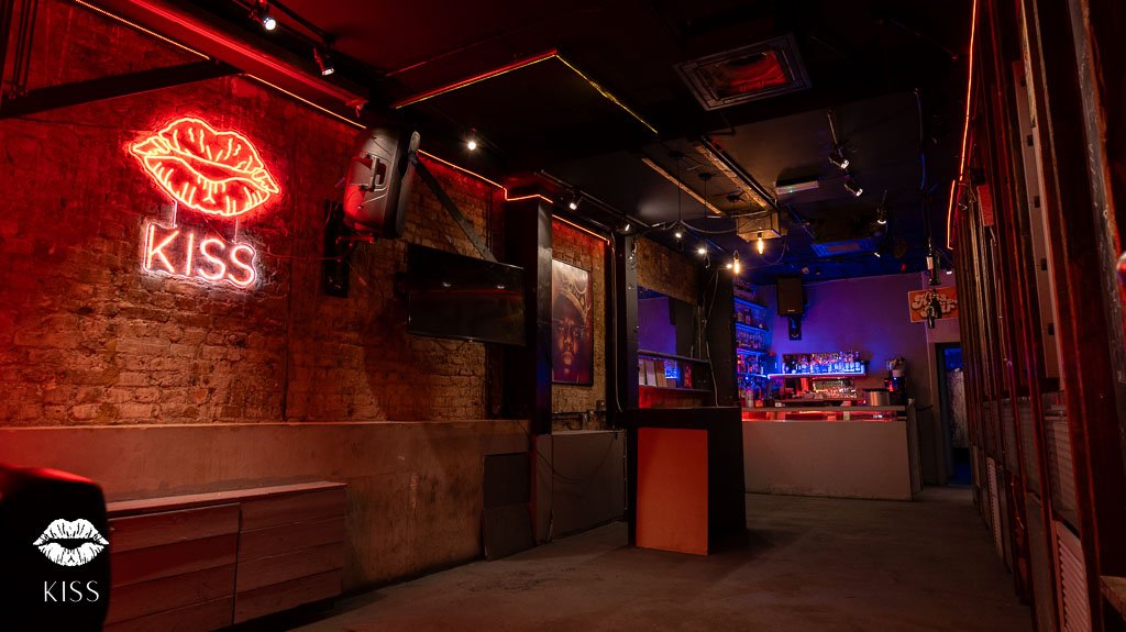 Dimly lit bar with exposed brick walls, neon sign of lips and the word 'KISS', bar counter with liquor bottles, musical equipment, and seating area.