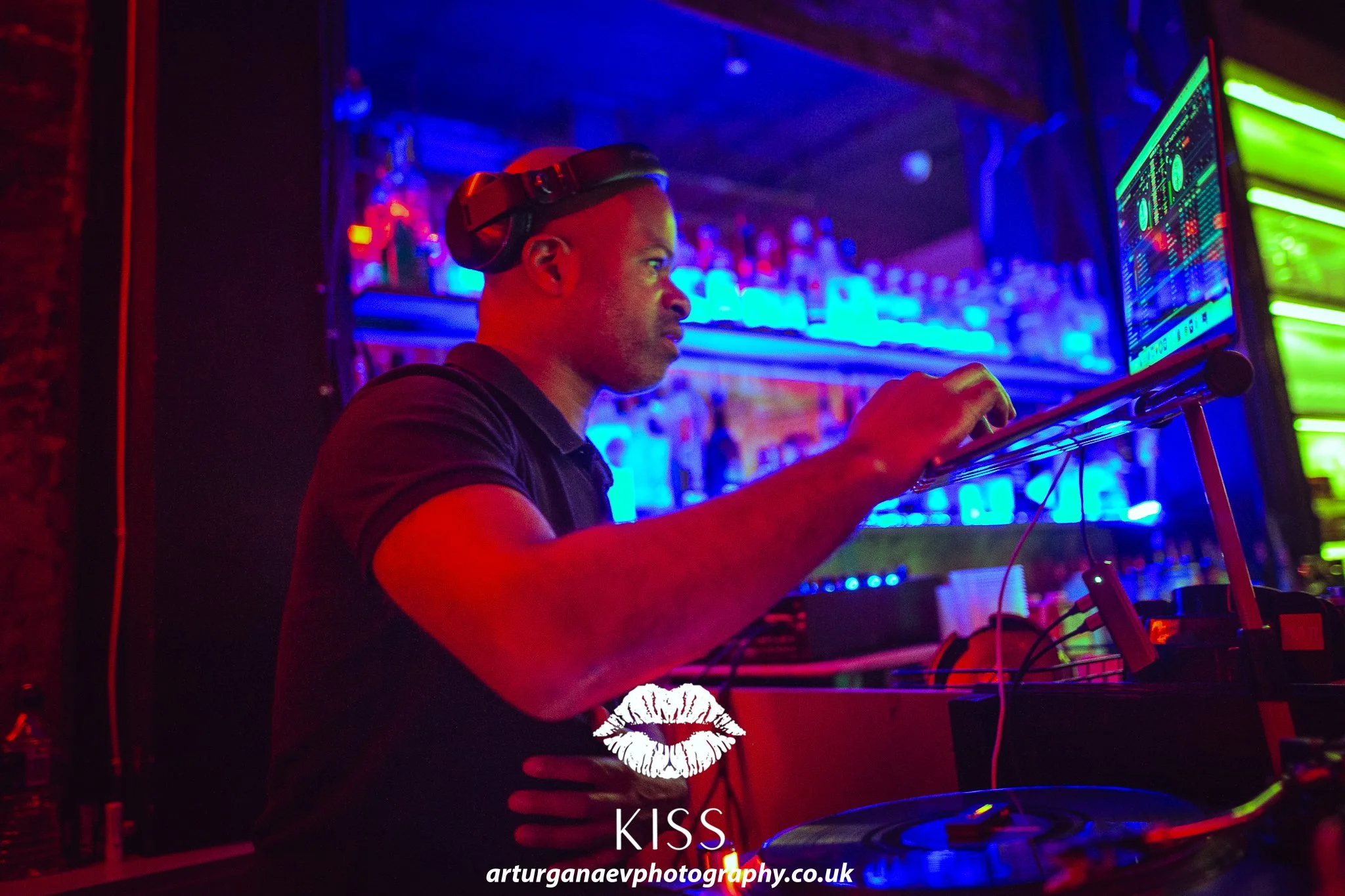 A DJ in a black shirt, wearing headphones, intensely focused on his turntable at a nightclub with colorful neon lights, a bar with bottles in the background, and a computer screen with DJ equipment.