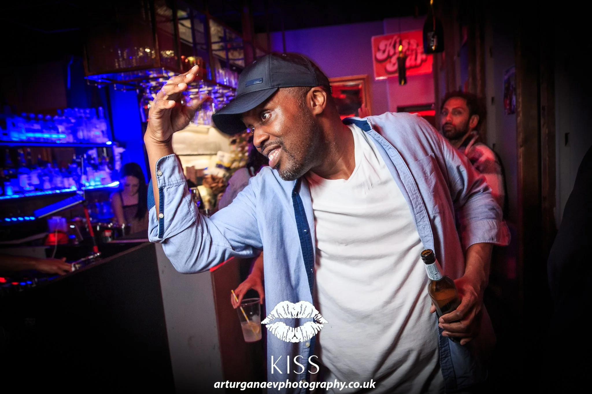 A man in a baseball cap, white t-shirt, and blue jacket holding a beer bottle in a dimly lit bar or club, dancing with a drink in his other hand. Neon and colorful lights in the background.