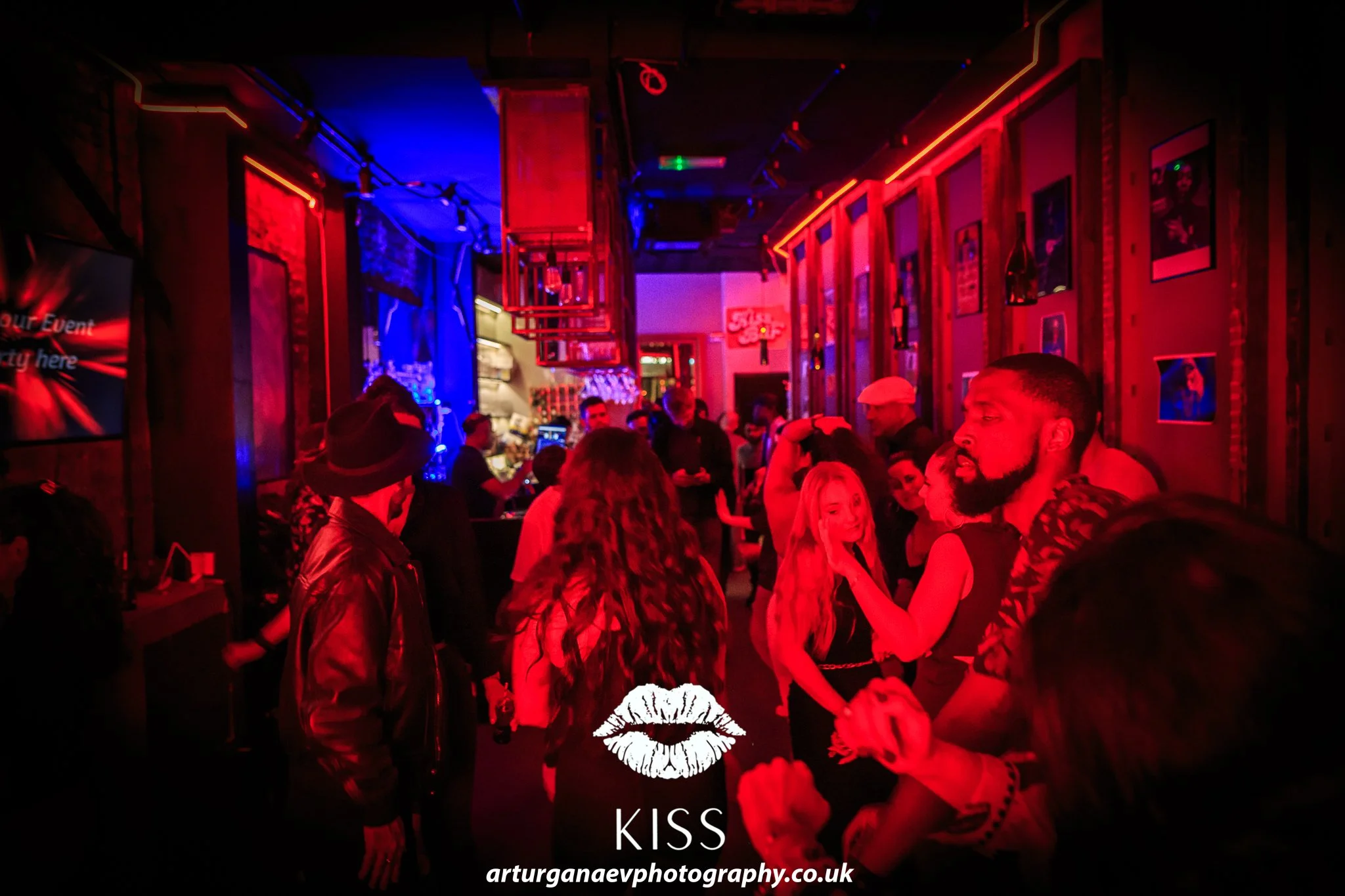 People dancing and socializing in a dimly lit bar with red and blue neon lights, framed pictures on the wall, and a visible bar area in the background.