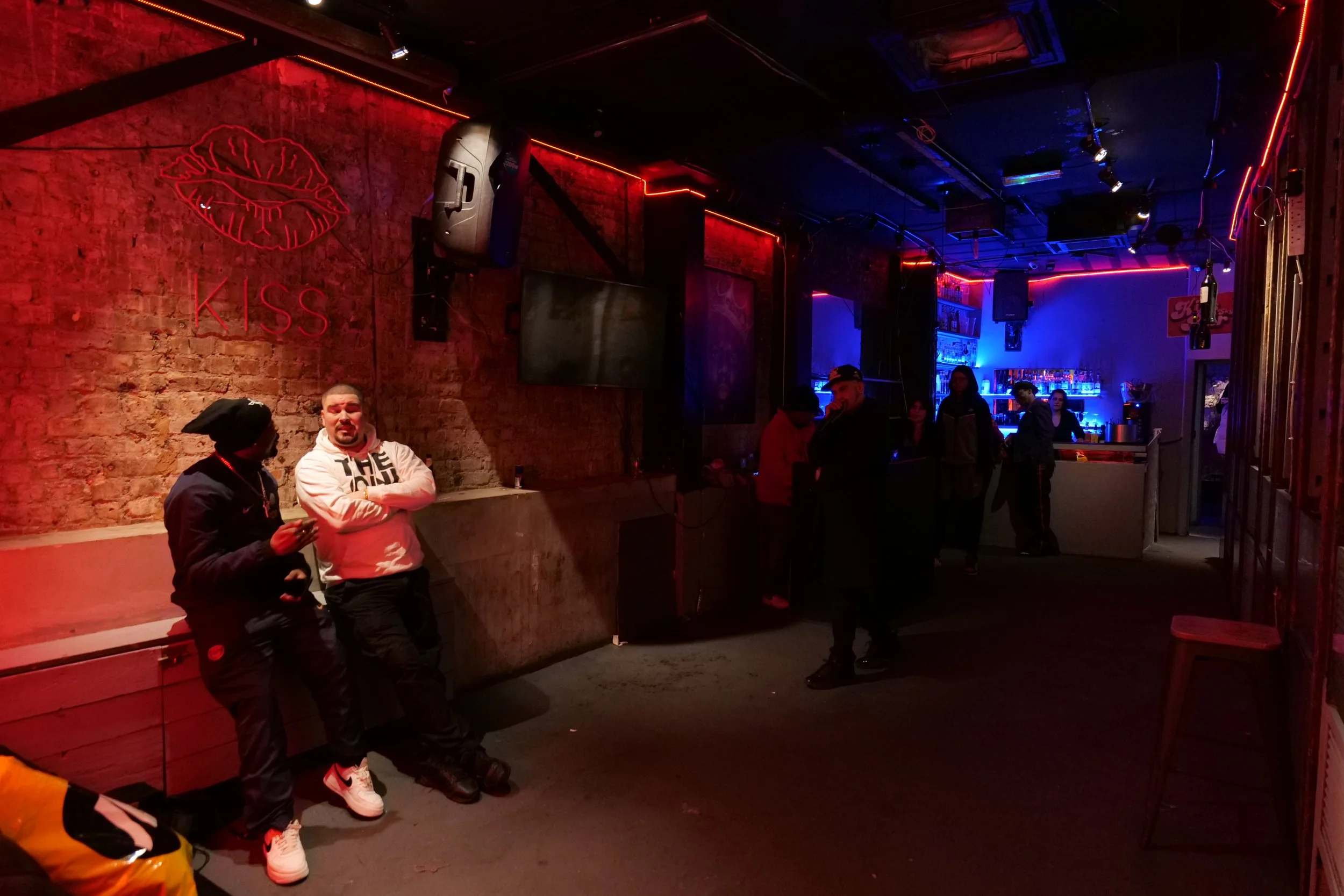 Interior of a dimly lit bar with a neon sign of red lips and the word 'KISS' on a brick wall, several people socializing near the bar area, illuminated by red and blue neon lighting.