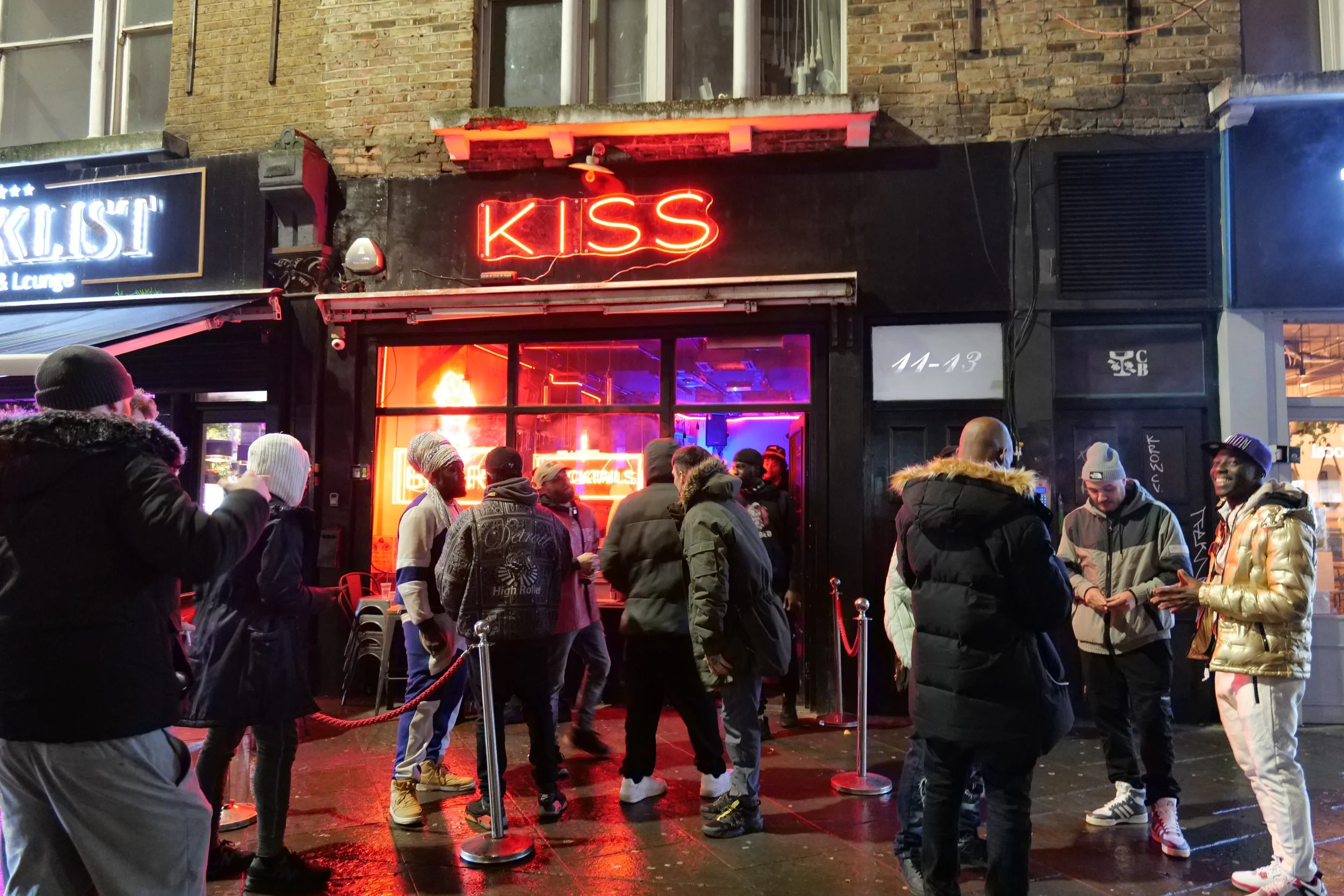A group of people waiting outside a nightclub called 'KISS' at night. The club has a neon red sign and a brightly lit interior visible through glass doors. The people are dressed warmly, indicating cold weather, and are standing in a line behind red 